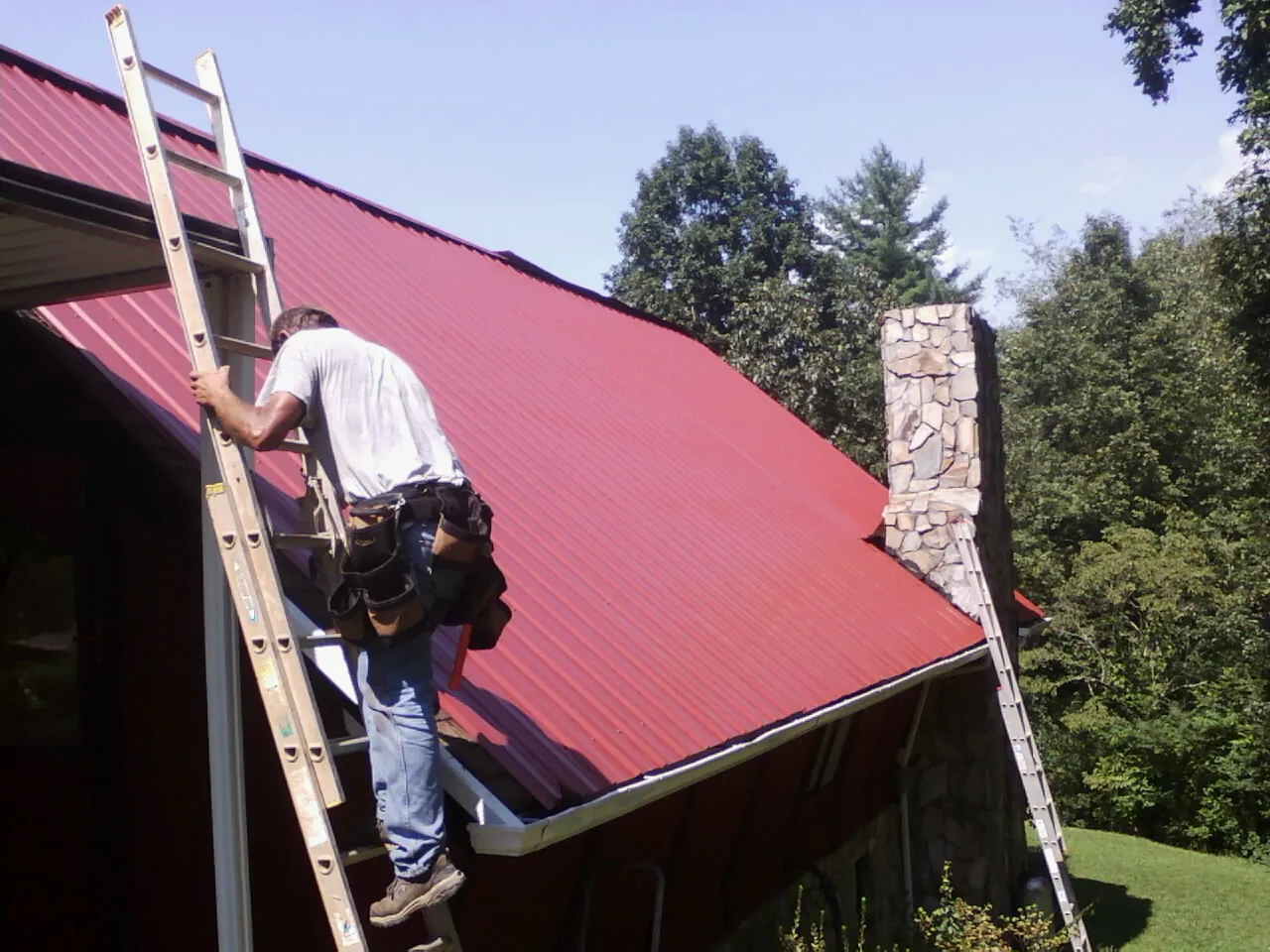 Roofer on a ladder beside a red metal roof and stone chimney — Soffit & Fascia Repair by Roofing Brothers in Perrysville
